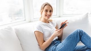 Digitale Services per App Junge Frau in Jeans und weißem T-Shirt sitzt auf einer hellen Couch und hat ein Smartphone in der Hand