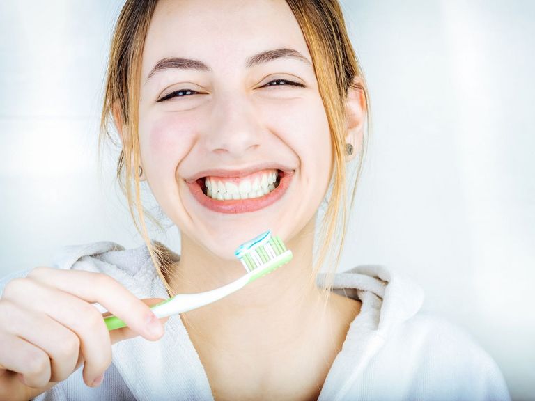 Lachende Junge Frau zeigt gepflegte Zähne und eine Zahnbürste mit Zahnpasta darauf