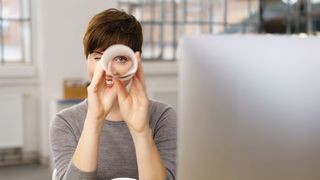 Stellenangebote Frau sitzt vor einem PC-Bildschirm und schaut suchend durch eine Papierrolle wie durch ein Fernrohr