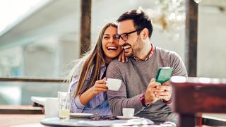 Paar sitzt mit Smartphone und Kaffee lachend an einem Tisch