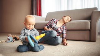 Burnout bei Müttern Baby spielt im Vordergrund und die Mutter sitzt erschöpft auf dem Boden und lehnt am Sofa