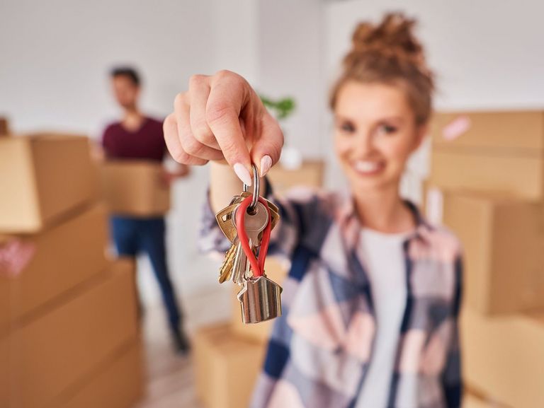 Junge Frau steht in einer Wohnung mit Umzugskartons und zeigt den Schlüssel direkt in die Kamera