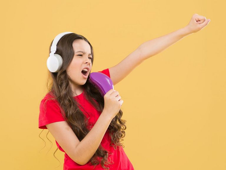 Junges Mädchen mit weißen Kopfhörern singt in eine Haarbürste und streckt dabei eine Faust in die Luft