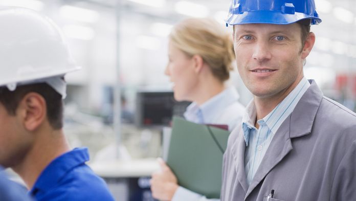 Betriebliches Gesundheitsmanagement Mitarbeiter in Fertigungshalle schaut in die Kamera