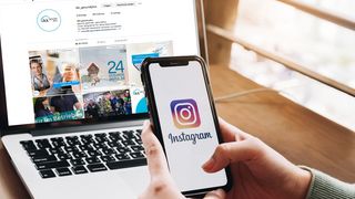 Instagram: Wir sind online! Hände halten ein Smartphone auf dem das Instagram-Logo zu sehen ist, im Hintergrund ein Laptop