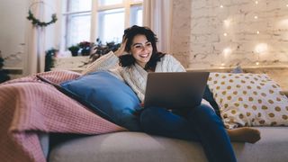 Frau sitzt mit einem Laptop auf den Knien auf einer Couch und im Hintergrund leuchten Adventslichter