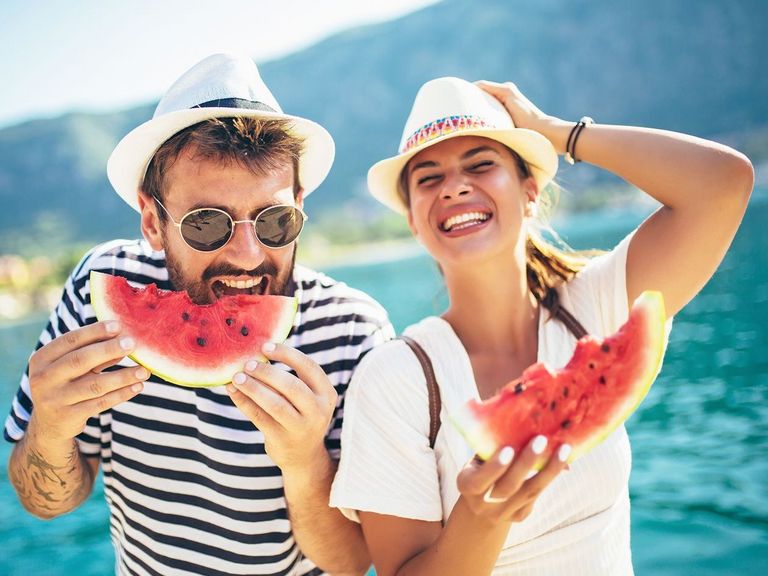 Mann und Frau vor einem See stehend, die lachend eine Melone essen