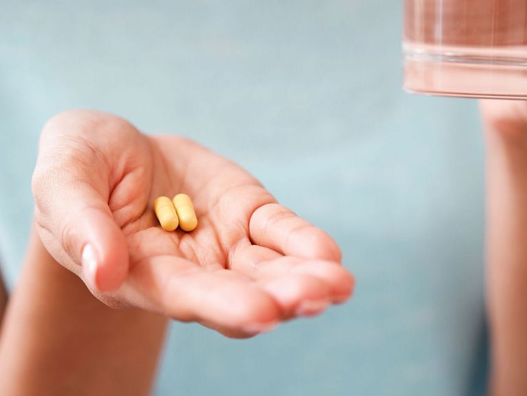 Frauenhand hält zwei gelbliche Medikamentenkapseln und im Hintergrund ist ein Glas Wasser zu sehen