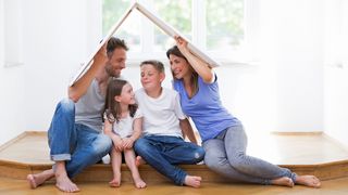 Familie sitzt glücklich in einer neuen Wohnung und hält sich symbolisch ein Pappdach über die Köpfe