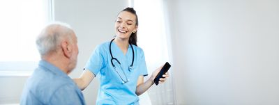 Pflegekraft in blauer Arbeitskleidung erklärt einem Pflegebedürftigen die Anwendung einer App auf dem Smartphone