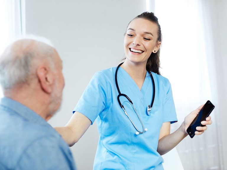 Pflegekraft in blauer Arbeitskleidung erklärt einem Pflegebedürftigen die Anwendung einer App auf dem Smartphone
