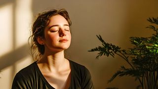 Mental Health Week Junge Frau sitzt meditierend in der Sonne, die durch ein Fenster hereinscheint