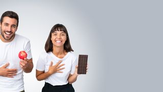 Anti-Diät-Tag Mann hält lachend einen roten Apfel in der Hand und die Frau hält eine Tafel Schokolade