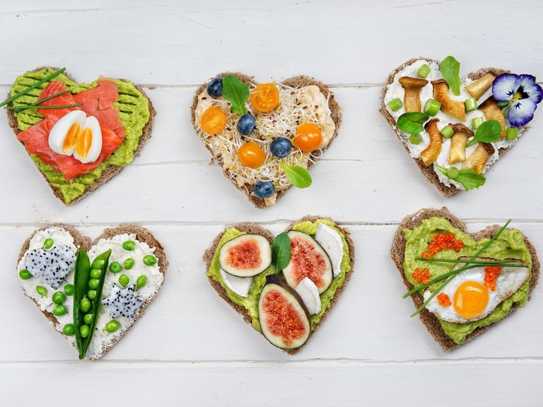 Vier bunt und gesund belegte Vollkornbrotscheiben in Herzform auf einem hell gestrichenem Holztisch