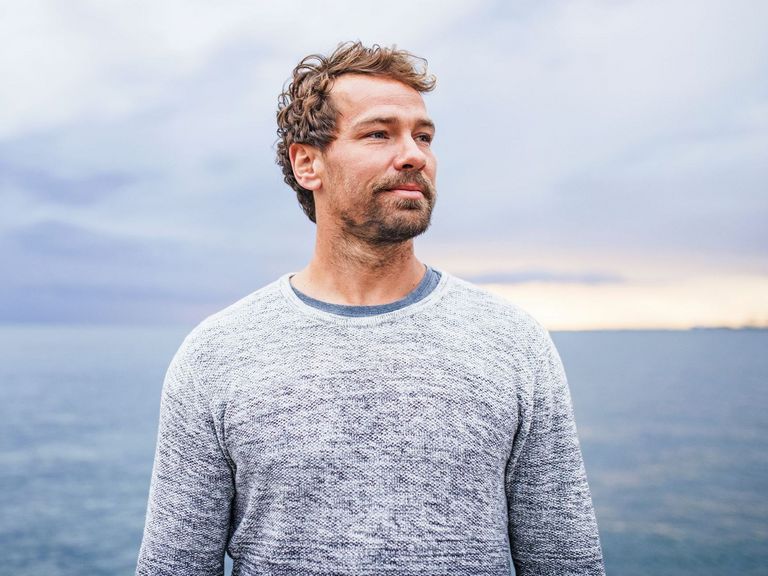 Mann schaut an einem Strand stehend an der Kamera vorbei