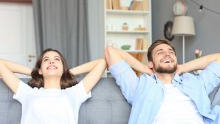 Pärchen sitzt mit hinter dem Kopf gelegten Händen entspannt und glücklich strahlend auf einer Couch