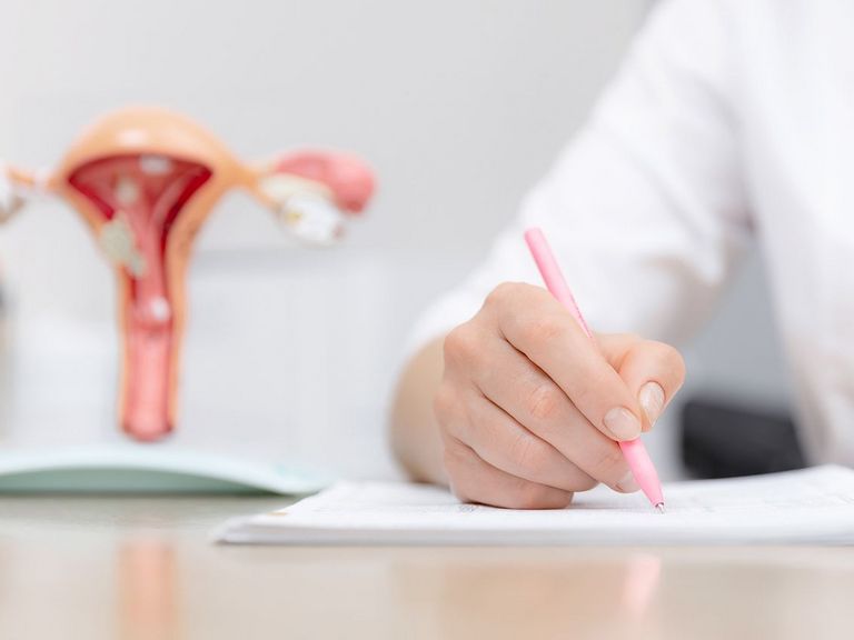 Frauärztin sitzt am Schreibtisch und notiert etwas, im Hintergrund ist ein Modell der Gebärmutter zu sehen