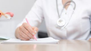 Gebärmutterhalskrebs Frauärztin sitzt am Schreibtisch und notiert etwas, im Hintergrund ist ein Modell der Gebärmutter zu sehen