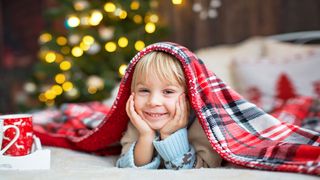 Hygge Kleiner, grinsender Junge mit einer karierten Decke auf dem Kopf und im Hintergrund leuchten Lichter