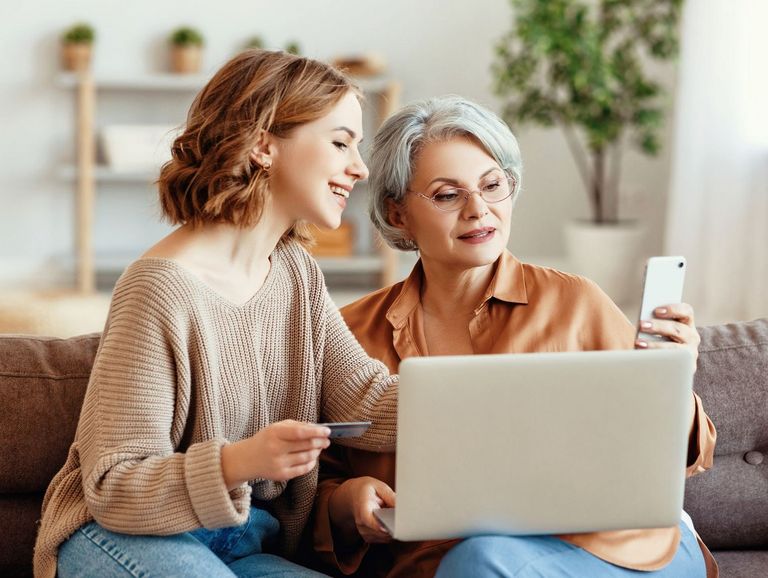 Zwei Frauen sitzen im Wohnzimmer auf einem Sofa und nutzen digitale Angebote per Laptop und Smartphone