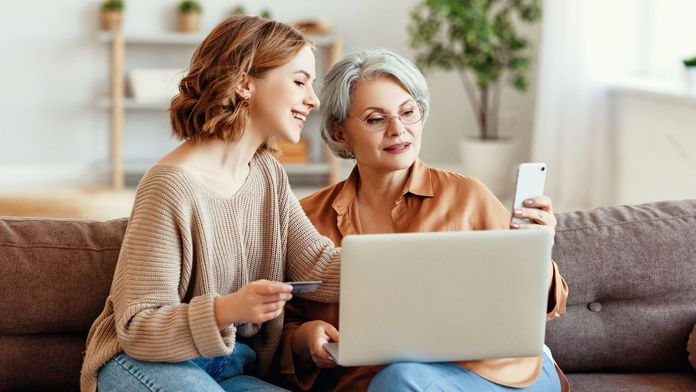 Zwei Frauen sitzen im Wohnzimmer auf einem Sofa und nutzen digitale Angebote per Laptop und Smartphone