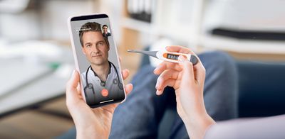 Hände halten ein Fieberthermometer und ein Smartphone, auf dem ein Videochat mit einem Arzt zu sehen ist