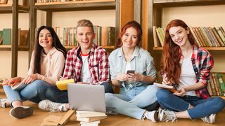Vier Teenager sitzen im Schneidersitz auf dem Boden vor einem großen Bücherregal und schauen in Bücher und in einen Laptop
