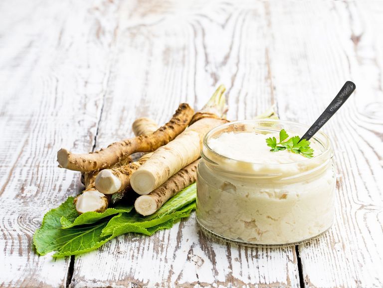 Mehrere kleine Meerrettichwurzeln und ein geöffnetes Schraubglas mit geriebenen Meerrettich liegen auf einem hellen Holztisch
