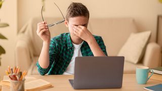 Teenager sitzt vor dem Laptop und reibt sich mit einer Hand die Augen