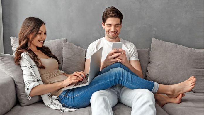 Das IKK-WebCenter Pärchen sitzt fröhlich mit Smartphone und Laptop auf einem grauen Sofa