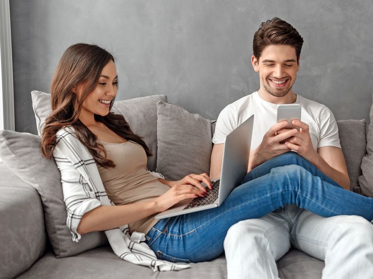 Pärchen sitzt fröhlich mit Smartphone und Laptop auf einem grauen Sofa