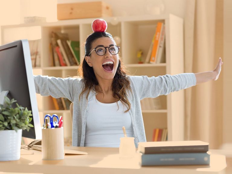 Dunkelhaarige Jugendliche sitzt am Schreibtisch und balanciert, mit seitlich ausgestreckten Armen, einen Apfel auf dem Kopf