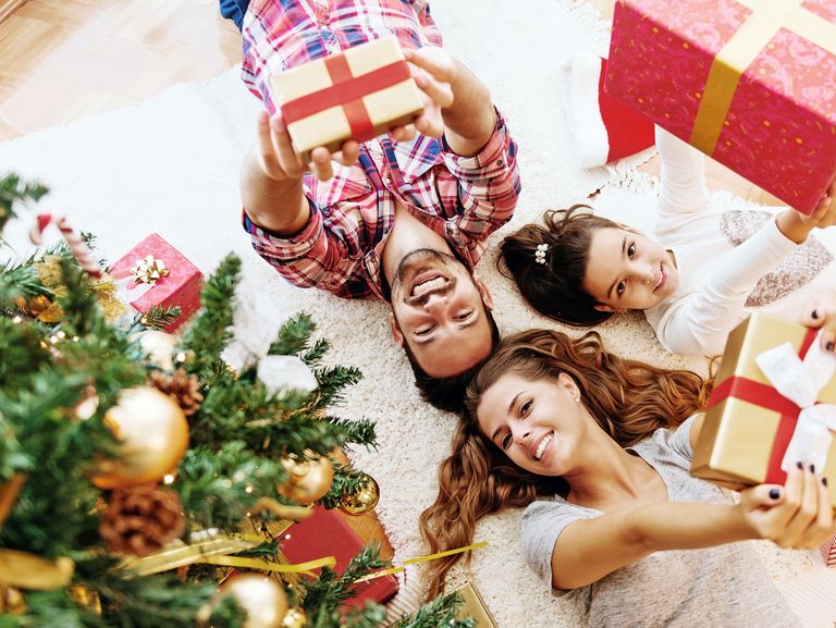 Familie liegt fröhlich unter einem Weihnachtsbaum und streckt Geschenke in die Höhe in Richtung Kamera