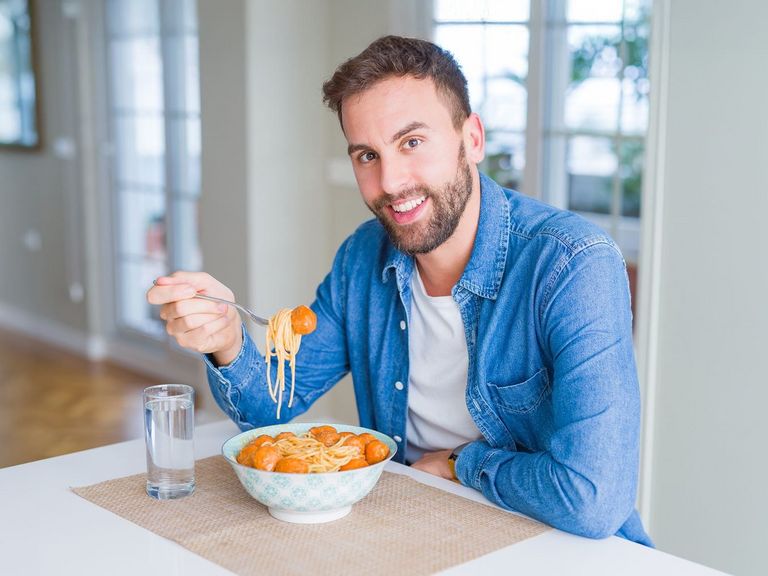 Mann mit Bart siitzt in der Küche beim Mittagessen und auf dem Tisch steht ein Glass mit Wasser