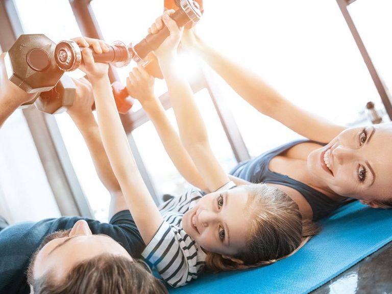 Dreiköpfige Familie liegt auf einer Turnmatte und trainiert mit Hanteln in der Luft