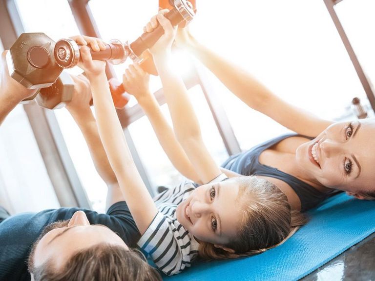 Dreiköpfige Familie liegt auf einer Turnmatte und trainiert mit Hanteln in der Luft