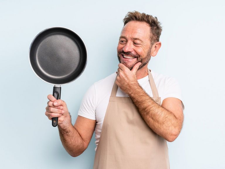 Mann mit beigefarbener Schürze hält eine Pfanne hoch und schaut verschmitzt