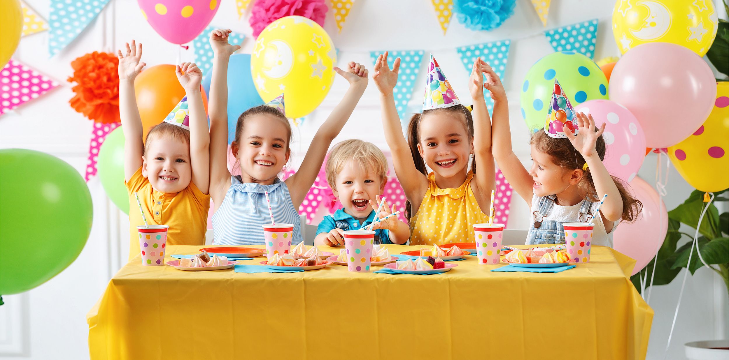 Fünf kleine Kinder jubeln mit erhobenen Armen inmitten bunter Geburtstagsdekoration