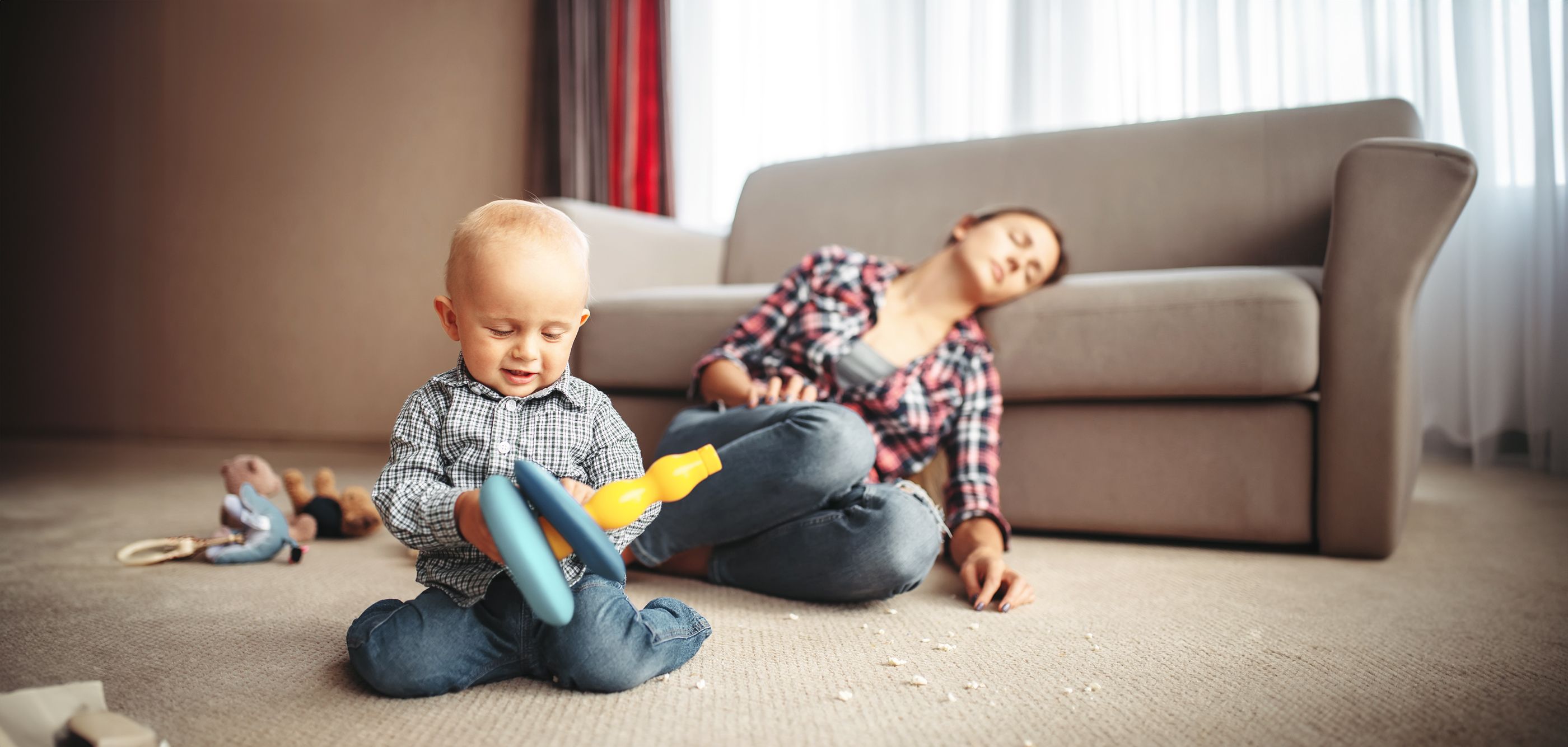 Burnout bei Müttern Baby spielt im Vordergrund und die Mutter sitzt erschöpft auf dem Boden und lehnt am Sofa