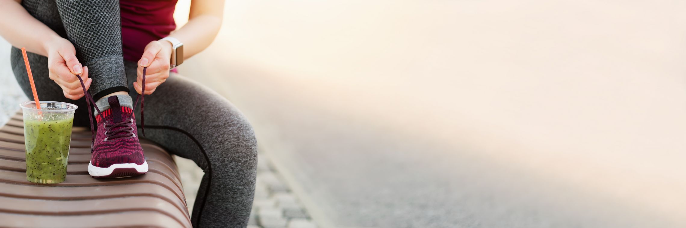 Sportlich gekleidete Frau sitzt auf einer Bank und bindet sich ihre Trainingsschuhe zu