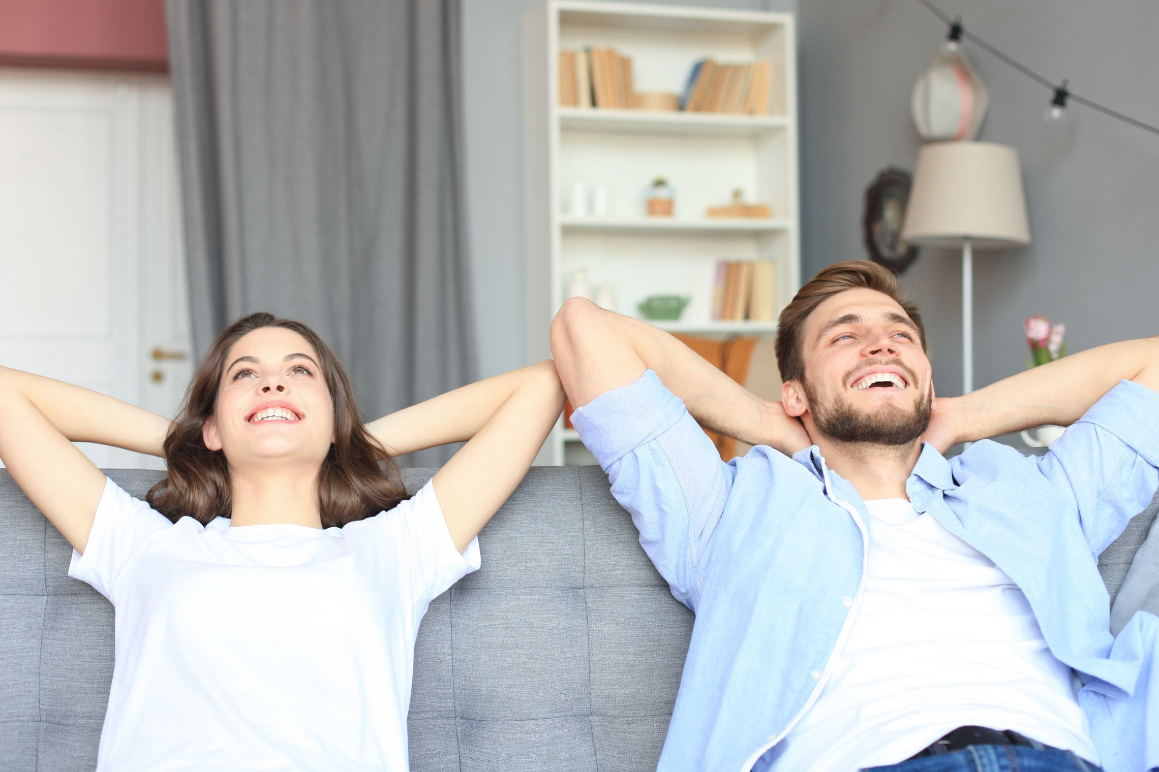 Pärchen sitzt mit hinter dem Kopf gelegten Händen entspannt und glücklich strahlend auf einer Couch