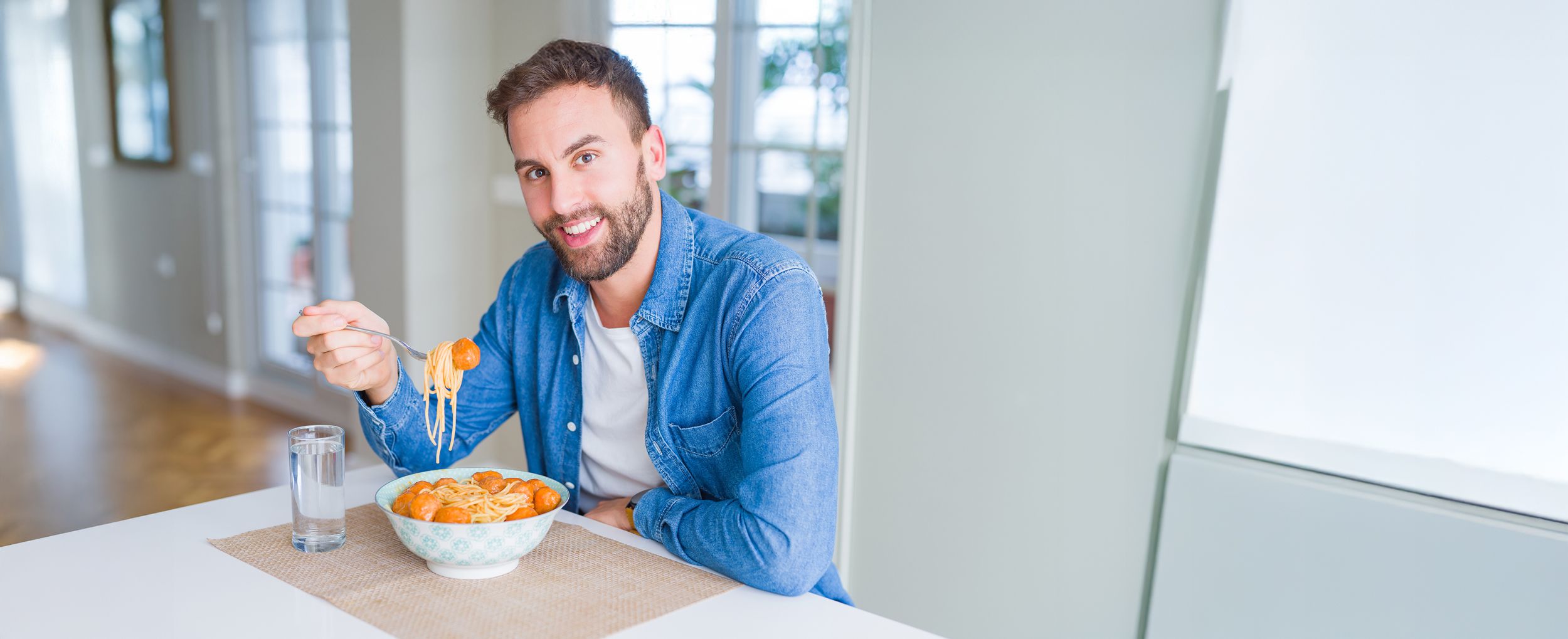 Mann mit Bart siitzt in der Küche beim Mittagessen und auf dem Tisch steht ein Glass mit Wasser