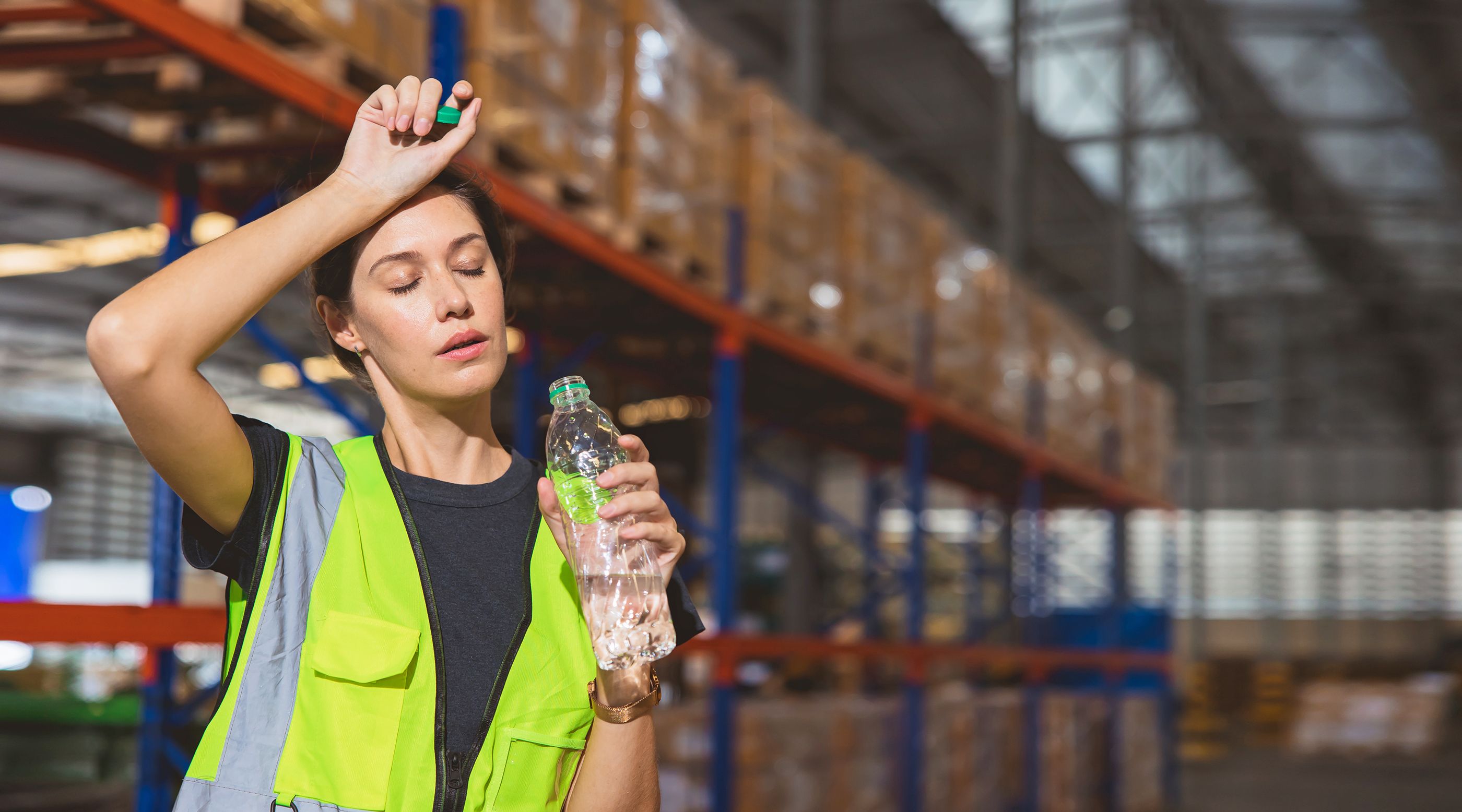 Lageristin ist erschöpft von der Hitze am Arbeitsplatz und hält eine offene Wasserflasche in der Hand