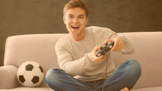 Teenager sitz im Schneidersitz mitfiebernd auf der Couch und hält eine Spieleconsole in seinen Händen, neben ihm liegt ein Fußball 