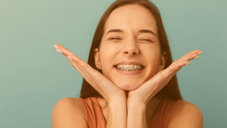 Brünette Jugendliche stützt ihr Kinn in ihre Handflächen und schenkt uns ein breites, mit einer Brackets-Zahnspange besetztes, Lächeln 