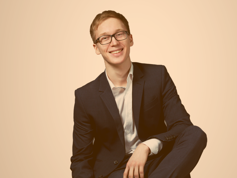 Jugendlicher, mit Brille und gepflegtem Haar, sitzt auf einem Stuhl und trägt einen blauen Anzug mit weißem Hemd darunter 