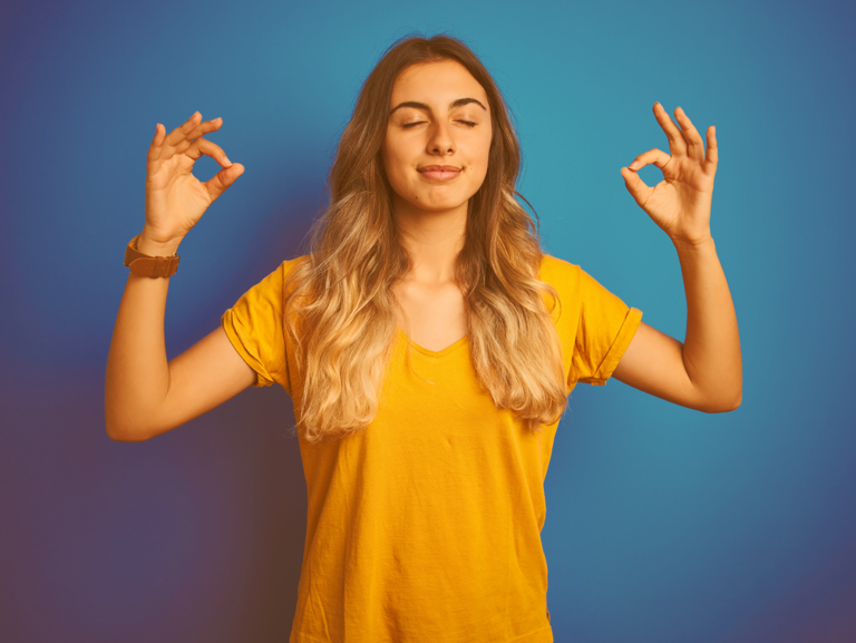 Junges Mädchen mit langen Haaren steh mit geschlossenen Augen und gehobenen Armen, während sie beidseitig das Gyan Mudra mit ihren Händen macht