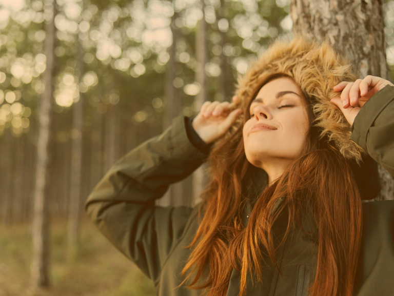 Jugendliche steht im Wald an eine Baum gelehnt und streckt ihr Gesicht lächelnd der winterlichen Sonne entgegen