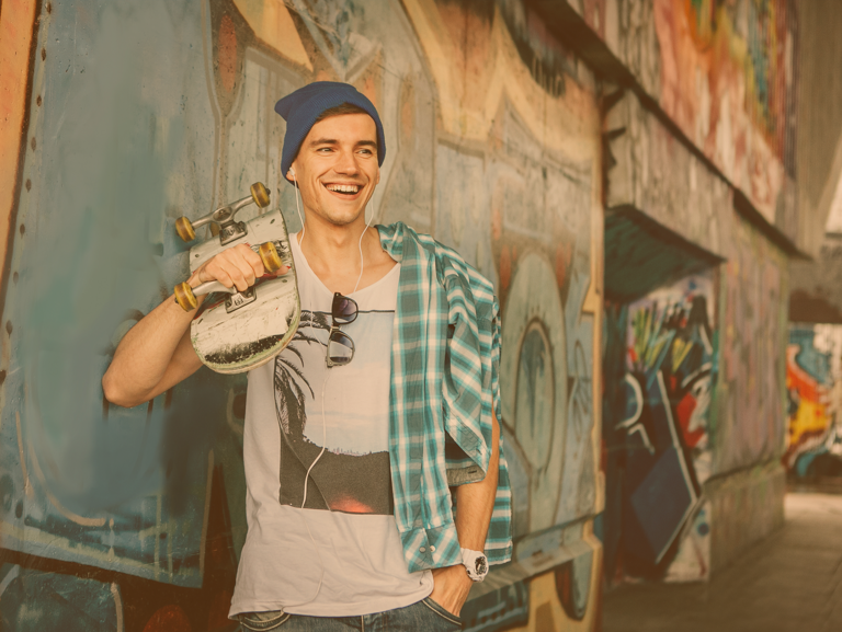 Junger Skater mit Hippster-Mütze steht mit Skateboard auf der Schulter an einer Graffiti-Wand gelehnt