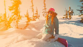 Jugendliche im Schnee-Outfit und verspiegelter Ski-Brille kniet mit Snowboard an den Füßen im Schnee zwischen verschneiter, sonniger Winterlandschaft